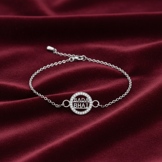 A silver chain bracelet with a circular pavé centerpiece engraved with 'BADA BHAi' in black lettering, displayed on a draped dark red velvet fabric.