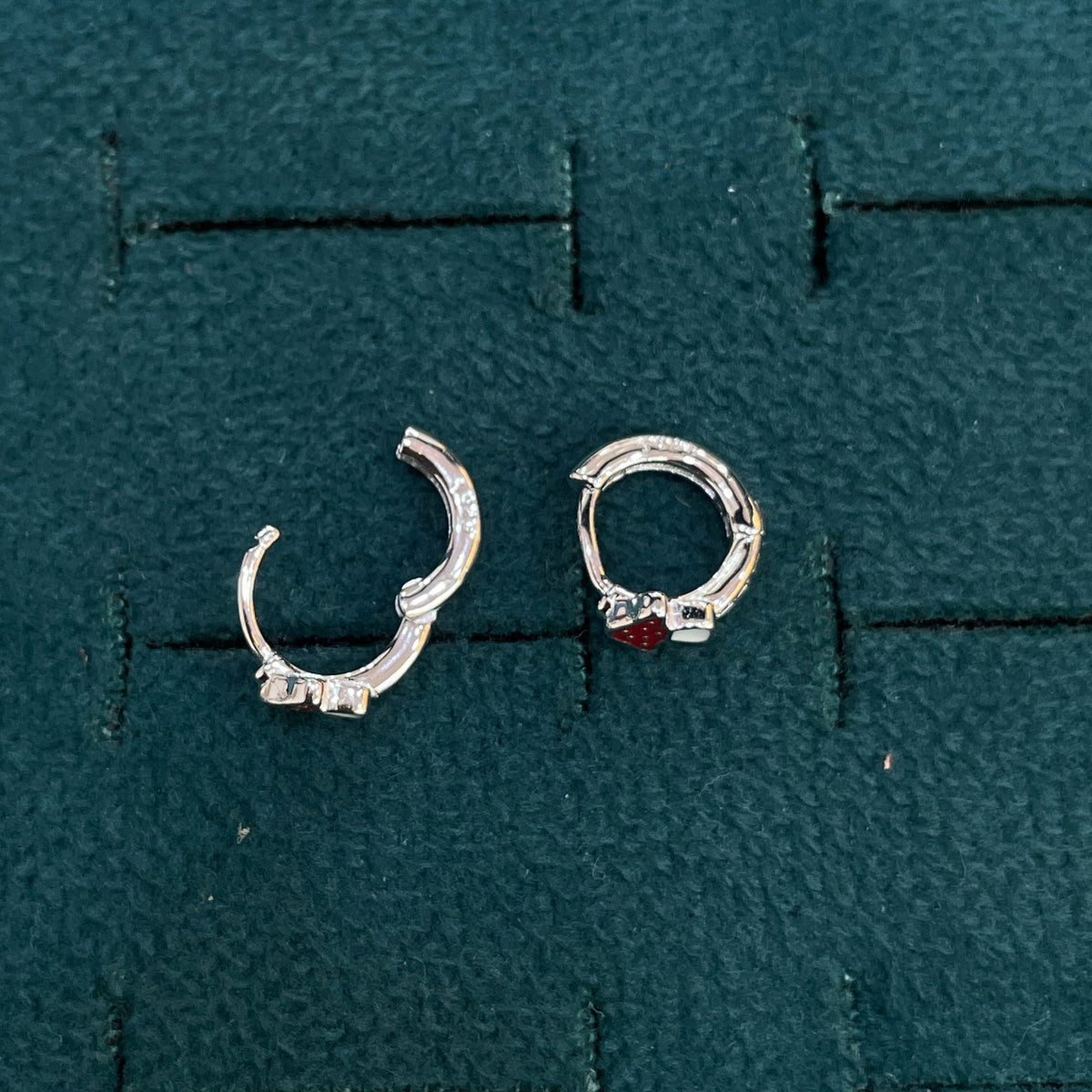 A pair of silver huggie hoop earrings displayed on a textured dark green jewelry display pad. One earring is plain silver, and the other features a small red gemstone accent.