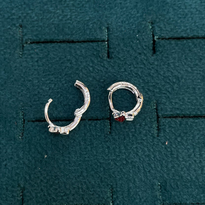 A pair of silver huggie hoop earrings displayed on a textured dark green jewelry display pad. One earring is plain silver, and the other features a small red gemstone accent.