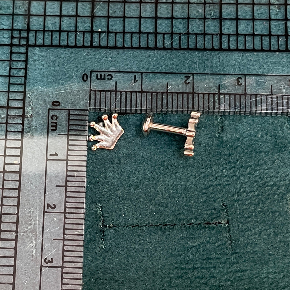 A pair of rose gold crown and anchor stud earrings placed on a dark green cutting mat next to clear plastic rulers for scale.