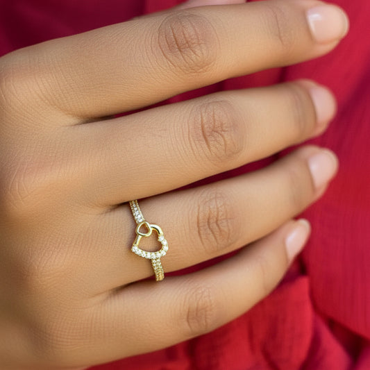 Gold ring with a heart design on a person's finger against a red fabric background