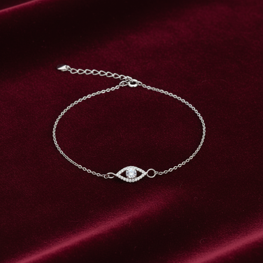 A delicate silver chain bracelet with an evil eye charm set with clear stones, displayed on a deep red velvet background.