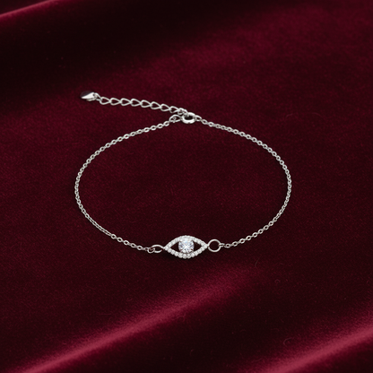 A delicate silver chain bracelet with an evil eye charm set with clear stones, displayed on a deep red velvet background.
