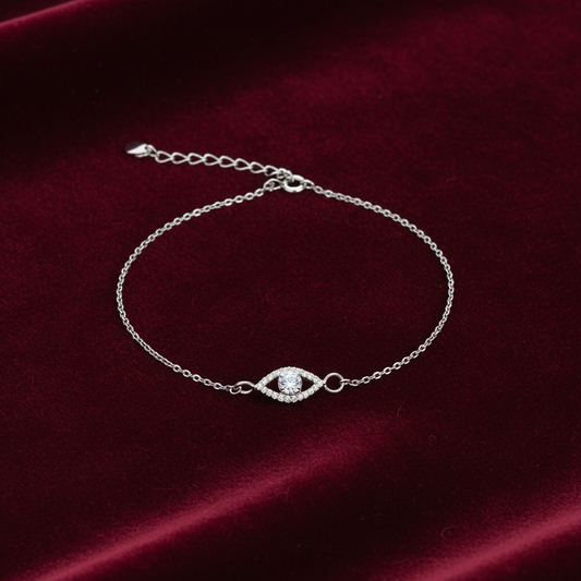 A delicate silver chain bracelet with an evil eye charm set with clear stones, displayed on a deep red velvet background.