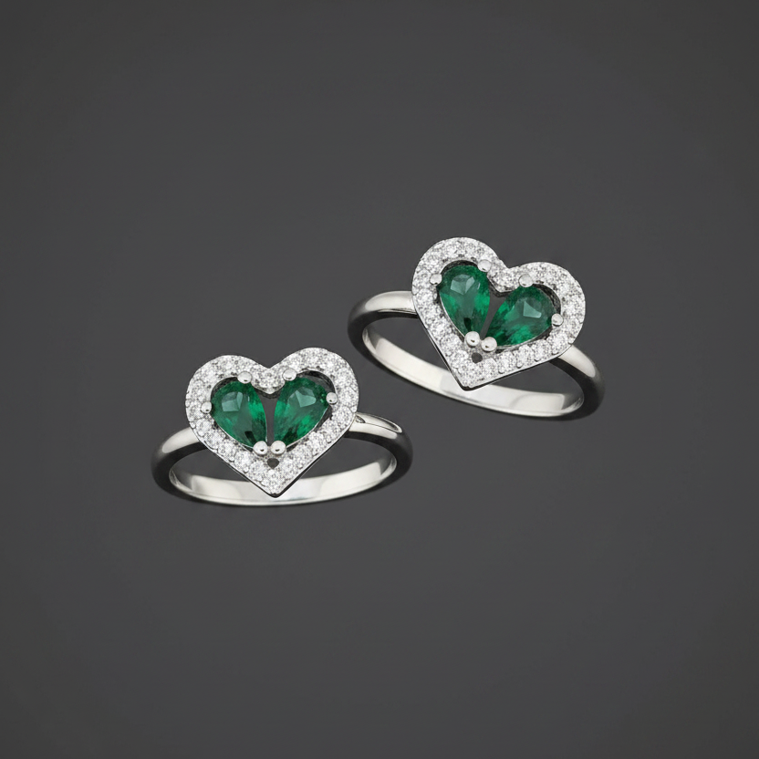 Heart-shaped rings with green gemstones on a wrist against a pink background