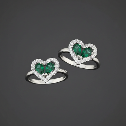 Heart-shaped rings with green gemstones on a wrist against a pink background
