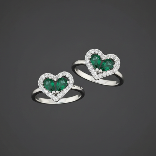 Heart-shaped rings with green gemstones on a wrist against a pink background