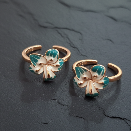 Butterfly-shaped gold and green rings on a dark stand with a pink background
